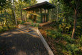 Exterior view of a modern ceramics studio situated in dense woodland, designed with a mono‑pitched roof, deep overhangs, and expansive multi‑panel glazing that frames immersive forest views. The green‑clad structure recedes quietly into the landscape, while an integrated wooden deck and concrete steps connect the light‑filled, highly functional interior workspace to its natural surroundings.