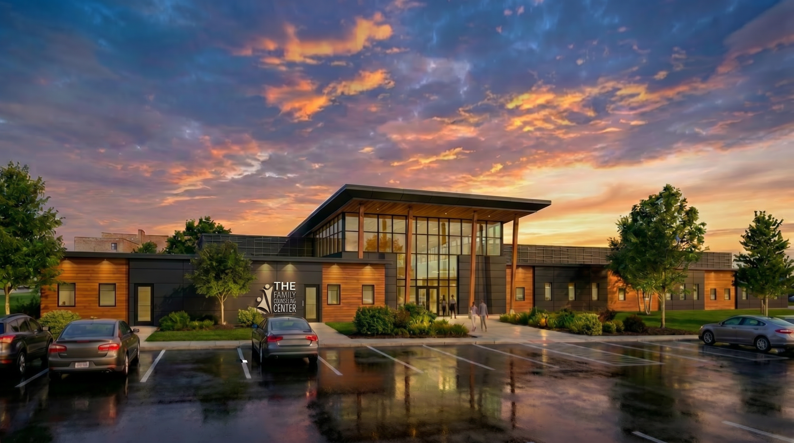 Exterior view of the Family Counseling Center designed with architecture+, showing a welcoming modern façade with tall glass panels, warm wood beams, and a clearly defined central entry. The building sits within landscaped greenery and is captured at sunset, with soft light reflecting across the wet pavement. The design emphasizes openness, safety, and human‑centered care through its transparent, approachable architectural expression.