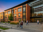 Exterior rendering of the proposed Roger Bacon Hall Addition at Siena University, designed with architecture+. The modern academic building features a mix of reddish‑brown brick and expansive glass façades, landscaped steps, benches, and ornamental grasses that frame active campus circulation. Captured at sunset with warm light reflecting across the pavement, the design emphasizes clean contemporary lines, transparent learning environments and welcoming connection points support interdisciplinary engagement