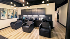 Interior view of the shampoo area at Alchemy Studios and Salon in Cohoes, NY, showing three black reclining wash chairs positioned before a counter with hair‑care products and neatly folded towels. Dark upper cabinetry contrasts with light walls, abstract blue‑and‑gold artwork, light wood flooring, and an exposed black ceiling with round pendant lighting, reinforcing the studio’s minimalist, light‑driven architectural aesthetic.