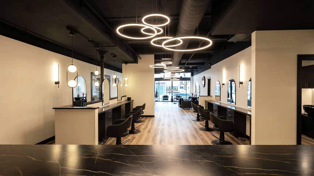 Interior view of the main styling floor at Alchemy Studios and Salon in Cohoes, NY, showing a long open layout with styling stations on both sides, black salon chairs, and large mirrors. Light wood flooring contrasts with the exposed black ceiling and circular pendant lighting, while natural light enters through the front windows, reinforcing the studio’s minimalist, light‑driven architectural concept.