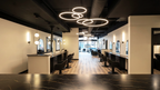 Interior view of the main styling floor at Alchemy Studios and Salon in Cohoes, NY, showing a long open layout with styling stations on both sides, black salon chairs, and large mirrors. Light wood flooring contrasts with the exposed black ceiling and circular pendant lighting, while natural light enters through the front windows, reinforcing the studio’s minimalist, light‑driven architectural concept.