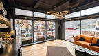 “Front‑of‑house lounge at Alchemy Studios and Salon in Cohoes, NY, showcasing floor‑to‑ceiling windows that flood the space with natural light. Black shelving displays curated products, while a tan leather sofa with black cushions anchors the seating area. Circular pendant lights, light wood flooring, and a dark ceiling reinforce the studio’s minimalist, light‑driven architectural aesthetic.