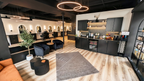 Interior view of the main styling floor at Alchemy Studios and Salon in Cohoes, NY, showing a row of illuminated styling stations, a waiting area with dark blue velvet chairs and a patterned rug, and a refreshment counter with black cabinetry, coffee machines, and product shelving. The light wood flooring and circular pendant lights reinforce the studio’s minimalist, light‑driven design concept.