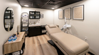 Interior view of a treatment room at Alchemy Studios and Salon in Cohoes, NY, designed with a minimalist palette and soft spatial transitions. The room includes a beige reclining treatment bed, black cabinetry with a sink, curated shelving, a large oval mirror, and three framed prints. Light wood flooring contrasts with the exposed black ceiling and recessed lighting, reinforcing the studio’s bright, gallery‑like aesthetic.